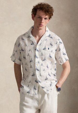 Jeune homme aux cheveux roux bouclés portant une chemise blanche à manches courtes avec des imprimés de bateaux à voile et de phares bleu marine, et un pantalon blanc.