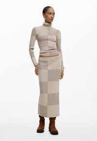 Conjunto de punto de dos piezas en beige claro con patrones acanalados verticales y horizontales. Cuenta con cuello alto y falda de longitud media, combinado con botas marrones hasta el tobillo.