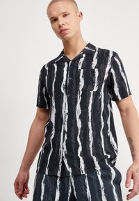 Camisa negra de manga corta con un patrón de rayas verticales blancas, cierre de botones en la parte delantera y un bolsillo de parche. Fabricada con una tela ligera y texturizada.