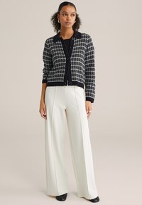 Sieviete, kas valkā melnu un baltu kvadrātiņiem rakstainu kardiganu ar melnu apkakli, platām gaiši krēmkrāsas biksēm un melnas kurpes, stāvot pret neitrālu fonu.