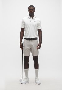 Άνδρας στέκεται κρατώντας μπαστούνι γκολφ, φορώντας λευκή πόλο, ανοιχτόχρωμο γκρι σορτς, λευκές κάλτσες και λευκά αθλητικά παπούτσια σε απλό φόντο.