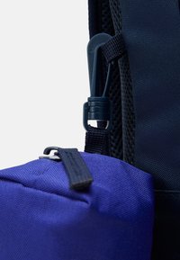 Pochette en tissu violet attachée à un sac à dos en mesh noir avec un mousqueton en plastique. Dotée d'une fermeture éclair et d'une surface texturée.