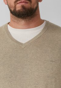 Beige V-Ausschnitt-Pullover mit weicher Textur und gerippten Bündchen. Mit einem dezenten Logo auf der Brust. Über einem weißen Rundhalshemd getragen.