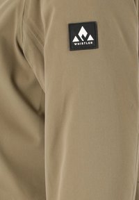 Khaki stofjakke med en glat tekstur, der har en sort rektangulær lap med et hvidt bjerglogo.
