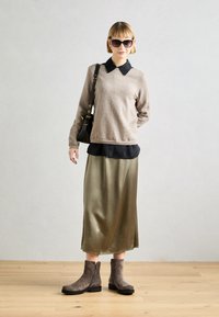 Suéter marrón claro sobre una camisa negra con cuello, falda satinada verde oliva y botines de ante grises. Se incluye un bolso negro y gafas de sol.