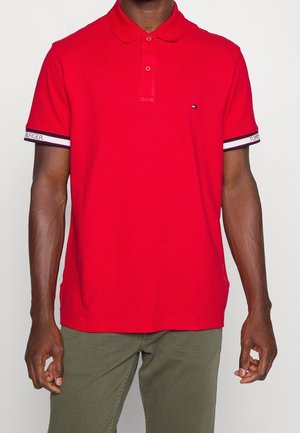 Mand iført rød poloshirt med to knapper og lille logo på brystet, logoer på ærmerne, kombineret med olivengrønne bukser.