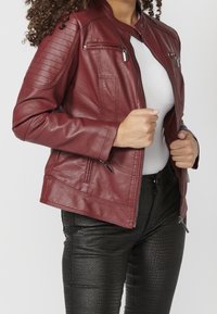 Burgundowa skórzana kurtka z stójką, żebrowymi akcentami na ramionach i zamykanymi kieszeniami, w połączeniu z czarnymi, strukturalnymi spodniami.