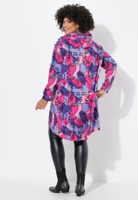 Tunic a maniche lunghe e multicolore con cappuccio. Presenta una stampa geometrica di volti in rosa, viola e blu su tessuto morbido. Leggings neri e stivaletti alla caviglia.