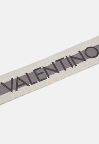 Cinta textil con "VALENTINO" en letras negras y en negrita sobre una franja gris, situada sobre un fondo claro de color beige texturizado.