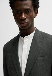 Blazer grigio a fantasia con rever a taglio, abbinato a una camicia bianca a collo alto. Tessuto liscio e dettagli sottili sulla tasca del blazer.