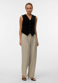 Gilet senza maniche in velluto nero con scollatura a V e cinque bottoni, abbinato a pantaloni palazzo beige a vita alta e décolleté neri con cinturini.