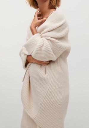 Vrouw gewikkeld in een zachte, roomkleurige geribbelde gebreide sjaal, met gekruiste armen en handen die haar nek en taille aanraken.