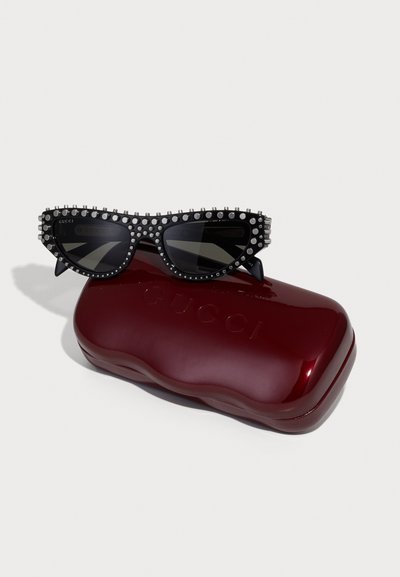 Occhiali da sole neri con punte affilate, dettagli borchiati e lenti scure, poggiati su un astuccio rosso lucido con la scritta "Gucci" in rilievo.