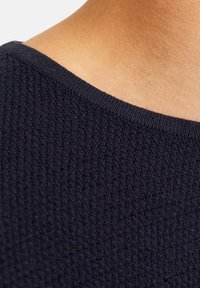 Nahaufnahme einer Schulter einer Person, die einen strukturierten, dunkelblauen Strickpullover mit ribbeligem Halsausschnitt trägt.