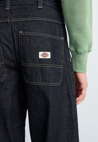 Sötét farmernadrág, egyenes szabású, sárga varrással és fehér Dickies logóval a hátsó zseben. Zöld pulóver látható.