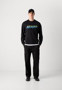 Man wearing black cap, black sweatshirt with gradient logo, black cargo pants, and black shoes, standing hands in pockets on white background.