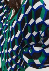 Kvinna i en grön knäppt kofta med en stickad tröja i blå, svart, grön och vit geometrisk kubmönster över.