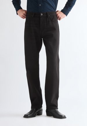 Personne portant un pantalon noir à jambes droites, des bottes noires à talons et une chemise sombre à manches longues, les mains sur les hanches.