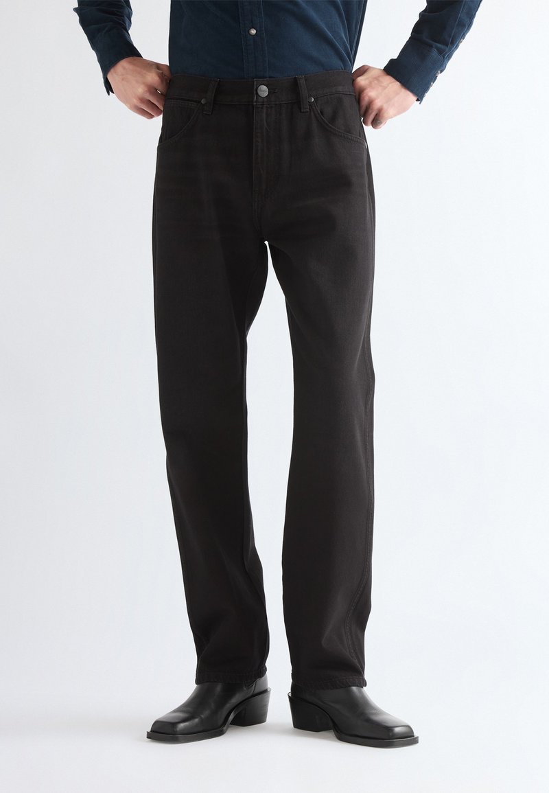 Personne portant un pantalon noir à jambes droites, des bottes noires à talons et une chemise sombre à manches longues, les mains sur les hanches.