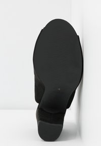 Chaussure à talons hauts en daim noir avec un bout arrondi et lisse et une semelle en caoutchouc texturée, affichant un petit accent doré près du talon.