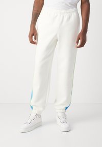 Hvide svedbukser med grønne og blå sidelister, elastisk talje og manchetter, parret med hvide sneakers. Glat stofstruktur.