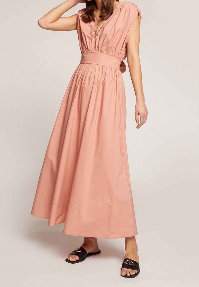 Femme portant une robe maxi sans manches rose avec une taille froncée et des sandales plates noires, se tenant devant un fond léger uni.