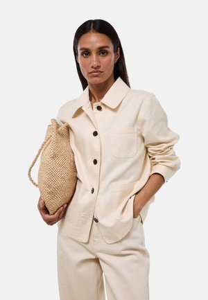Femme portant une veste beige boutonnée et un pantalon, tenant un sac tressé beige d'une main, l'autre main dans la poche, regardant devant.