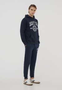 Felpa con cappuccio blu navy e pantaloni jogger con grafica "BOSTON", tasca frontale e polsini a costine. Sneakers bianche con strisce nere sui lati.