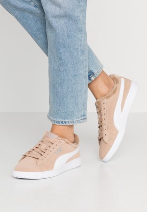 Fødder iført beige Puma-sneakers med hvide såler og pelsfor, kombineret med lyseblå opkørte jeans på en ensfarvet baggrund.