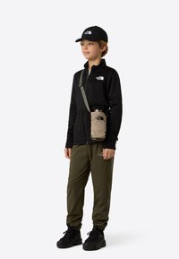 Black zip-up fleece jacket, olive green pants, black cap, and black boots; features a beige crossbody bag with a water bottle.