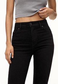 Jean noir taille haute avec une coupe classique à cinq poches, fermeture par bouton en métal et détails de couture subtils. Le tissu présente une texture lisse.