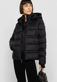 Zwarte donzen jas met capuchon, doorgestikt ontwerp, ritssluiting aan de voorkant en zijzakken. Soepele textuur met een logodetail op de mouw.