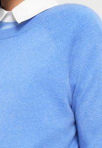 Pull bleu clair en maille fine, avec un col rond et des manches raglan. Texture lisse avec de légères variations.