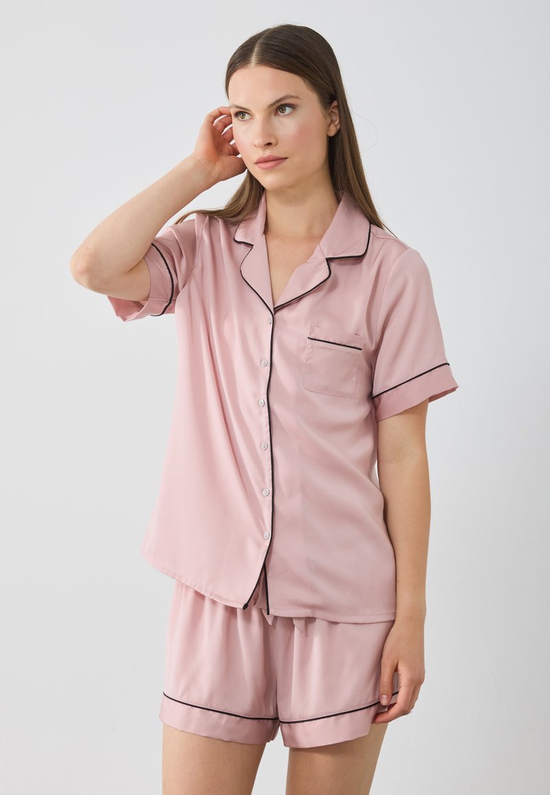 Korte roze satijnen pyjama set met zwarte piping, bestaande uit een korteknopschrift bovenkant met een borstzak en bijpassende shorts.