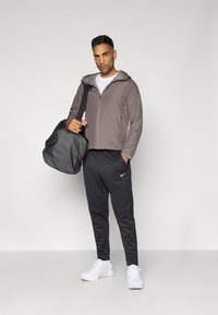 Grå zip-up jacka med huva, svarta träningsbyxor med vitt Nike-logotyp, och vita sneakers. Utrustad med en svart gymväska.
