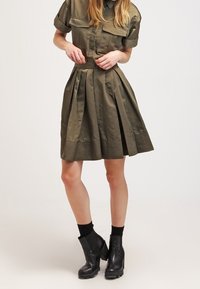 Olivinvihreä, lyhythihainen paitamekko, jossa laskostettu hameosa. Siinä on kaksi rintataskua, vyötäröllä vyö ja teksturoitu helma. Yhdistetty mustien nilkkureiden kanssa.