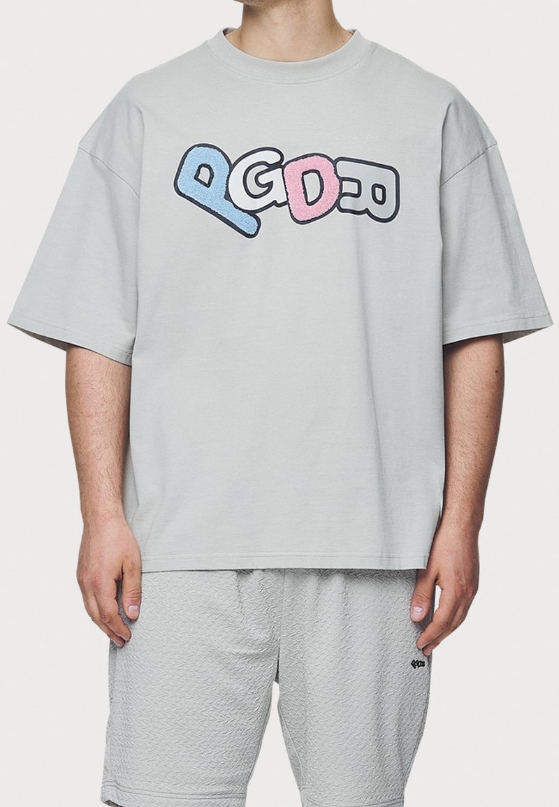 Camiseta de gran tamaño gris claro con mangas cortas, que presenta un diseño gráfico divertido en varios colores, en azul y rosa. Material de algodón, ajuste relajado.