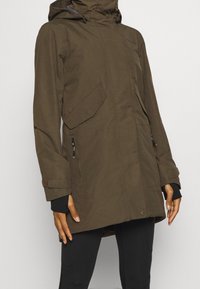 Olivgrön parka-jacka med huva, framknäppning med tryckknappar, fickor med dragkedja på sidorna, justerbara ärmslut och en slät yta.