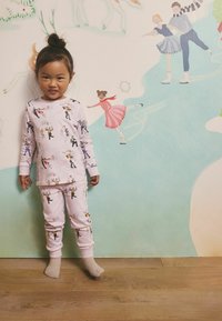 Šviesiai rožinė pižamos komplektas su vaikų šokių ir čiuožimo ledu vaizdu, su ribotomis rankogalių apdailomis ir minkšta tekstūra. Stovi ant medinių grindų.