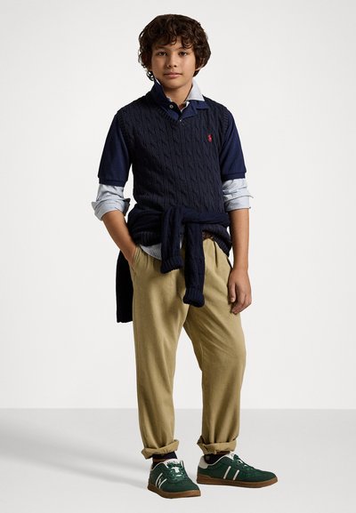 Ναυτικό πλεκτό γιλέκο πάνω από ένα μπλε πουκάμισο με γιακά, κρεμ παντελόνι και πράσινα αθλητικά παπούτσια με άσπρες ρίγες. Χαλαρή, στρώματη εμφάνιση με αναδιπλωμένα μανίκια.