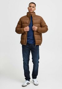 Brun pufferjakke med høy krage, glidelås og stikket design. Båret over en blå skjorte, kombinert med mørke jeans og hvite joggesko.
