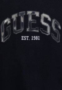 Černá mikina s vyvýšeným, lesklým bílým logem "GUESS" s maskáčovým vzorem a vyšitým "EST. 1981" pod ním.