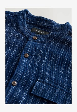 Sötétkék, texturált anyagból készült ing, függőleges fehér csíkokkal. Álló gallérral és barna gombokkal rendelkezik. Tartalmaz egy mellzsebet.