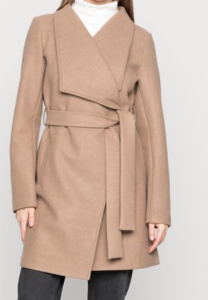 Femme portant un manteau en laine beige ceinturé sur un col roulé blanc, associé à un pantalon gris foncé, debout devant un fond clair uni.