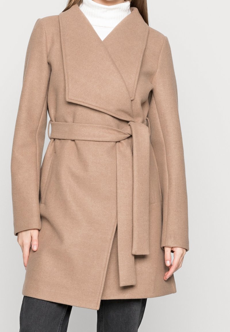 Femme portant un manteau en laine beige ceinturé sur un col roulé blanc, associé à un pantalon gris foncé, debout devant un fond clair uni.