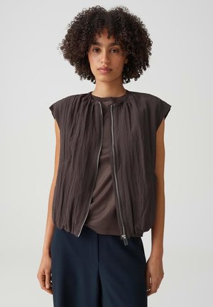 Jonge vrouw met krullend haar draagt een mouwloos donkerbruin vest met rits over een bruin shirt en marineblauwe broek, staand tegen een effen achtergrond.