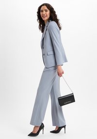 Frau mit lockigem Haar, die einen hellblauen Hosenanzug und schwarze High Heels trägt, hält eine schwarze Valentino-Handtasche mit einer silbernen Kettenschlaufe.