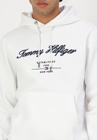 Hvit hettegensér med lomme foran, snorhette og brodert "Tommy Hilfiger"-logo i marineblått. Laget av mykt stoff.