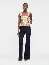 Femme portant un haut sans manches orné de dentelle fleurie et un jean évasé foncé taille haute avec des talons ouverts noirs sur un fond uni.