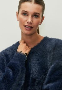 Maglione in pelliccia sintetica blu notte con maniche lunghe e finitura testurizzata, abbinato a un braccialetto con ciondoli argentati su un polso.
